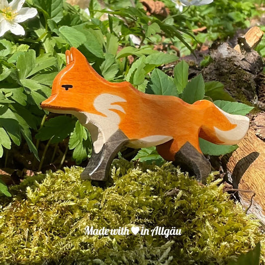 Handgefertigtes Holzspielzeug Fuchs Fähe rot von Allgäuer Holztiere, orange und schwarz bemalt mit pflanzlichen Farben, stehend in natürlicher Umgebung auf Waldboden oder Moos, aus bayerischem Ahornholz gefertigt, im Allgäu von Hand hergestellt und für Kinder ab 3 Jahren geeignet.