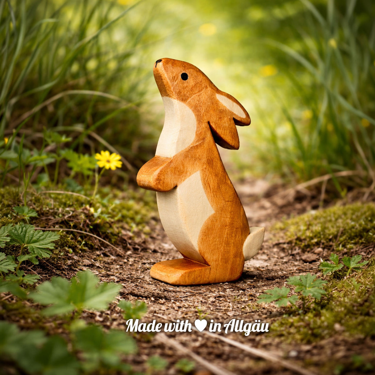 Handgefertigtes Holzspielzeug Feldhase Rammler Männchen von Allgäuer Holztiere, auf einem Waldweg zwischen Gras und kleinen Blumen stehend, im Hintergrund unscharfe grüne Vegetation, aus bayerischem Ahornholz gefertigt, mit pflanzlichen Farben braun und schwarz bemalt, im Allgäu produziert und für Kinder ab 3 Jahren geeignet.
