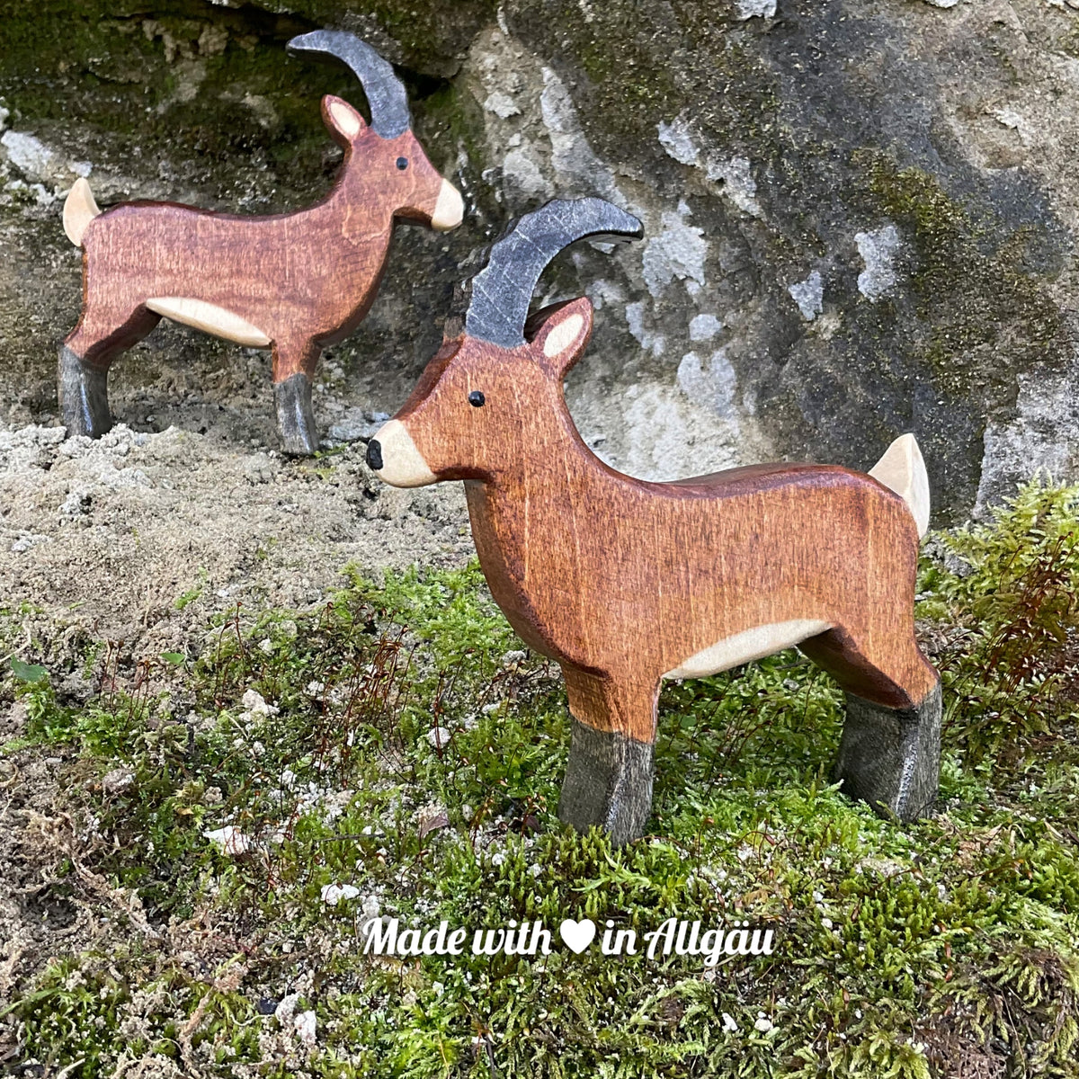 Handgefertigtes Holzspielzeug Steinbock Bock von Allgäuer Holztiere, stehend auf Moos und Felsen in natürlicher Berglandschaft der Alpen, aus bayerischem Ahornholz gefertigt, braun und schwarz bemalt mit pflanzlichen Farben, produziert im Allgäu und für Kinder ab 3 Jahren geeignet.