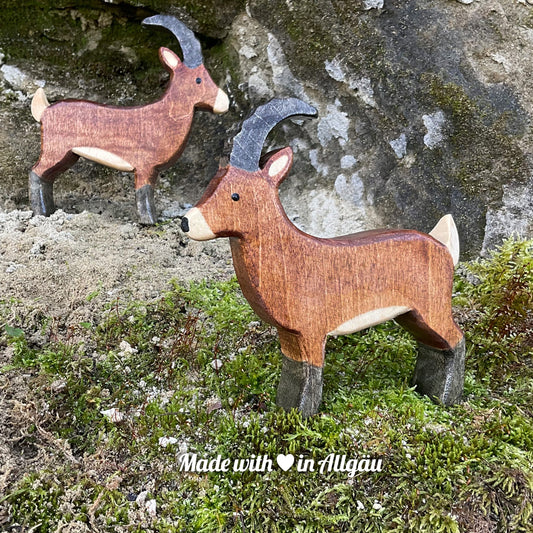 Handgefertigtes Holzspielzeug Steinbock Bock von Allgäuer Holztiere, stehend auf Moos und Felsen in natürlicher Berglandschaft der Alpen, aus bayerischem Ahornholz gefertigt, braun und schwarz bemalt mit pflanzlichen Farben, produziert im Allgäu und für Kinder ab 3 Jahren geeignet.