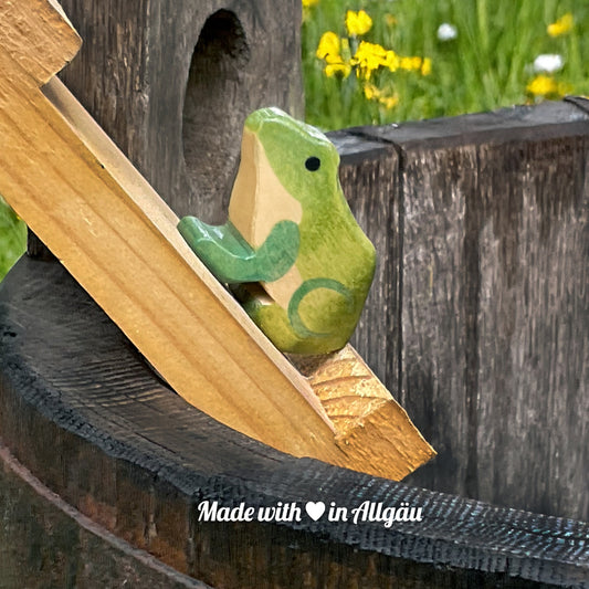 Handgefertigtes Holzspielzeug Frosch Weibchen von Allgäuer Holztiere, hellgrün und dunkelgrün bemalt mit pflanzlichen Farben, sitzend in Regentonne oder natürlicher Uferlandschaft, aus bayerischem Ahornholz gefertigt, im Allgäu von Hand hergestellt und für Kinder ab 3 Jahren geeignet.