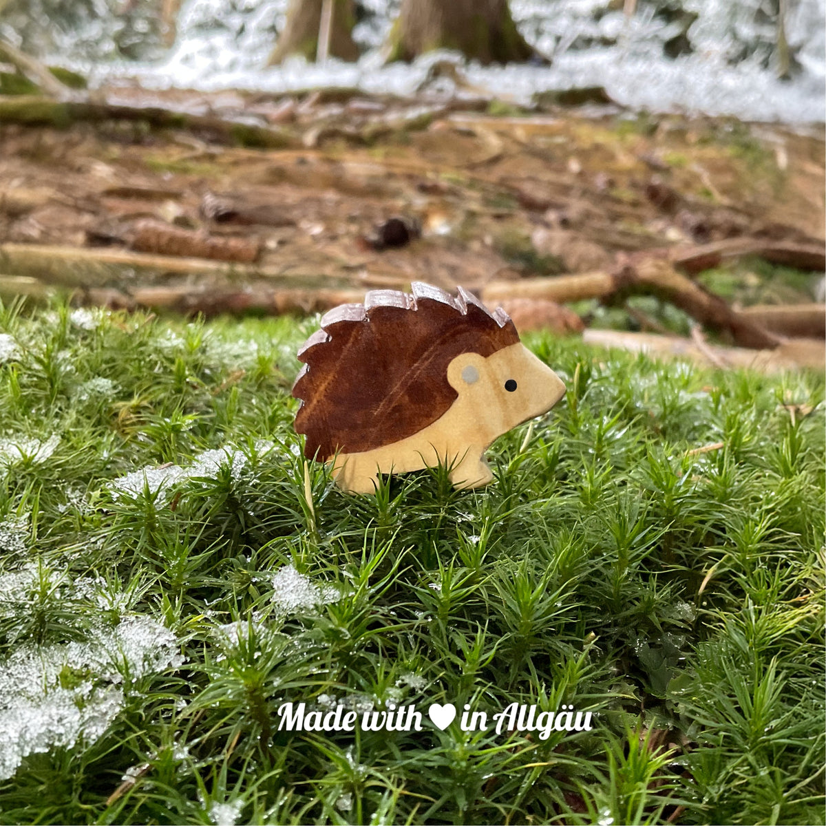 Handgefertigtes Holzspielzeug Igel Baby stehend von Allgäuer Holztiere, auf moosigem Waldboden mit etwas Schnee, im Hintergrund unscharfe Baumstämme, aus bayerischem Ahornholz gefertigt, braun und grau mit pflanzlichen Farben bemalt, im Allgäu produziert und für Kinder ab 3 Jahren geeignet.