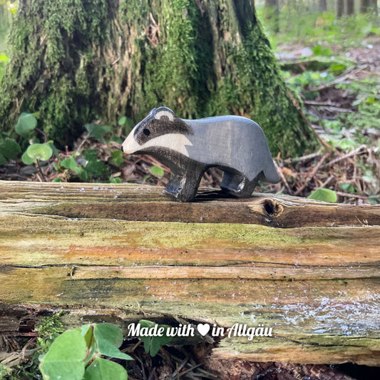 Handgefertigtes Holzspielzeug Dachs-Baby grau stehend von Allgäuer Holztiere, auf bemoostem, verwittertem Holz in natürlicher Waldlandschaft platziert, aus bayerischem Ahornholz gefertigt, grau und schwarz mit pflanzlichen Farben bemalt, im Allgäu produziert und für Kinder ab 3 Jahren geeignet.