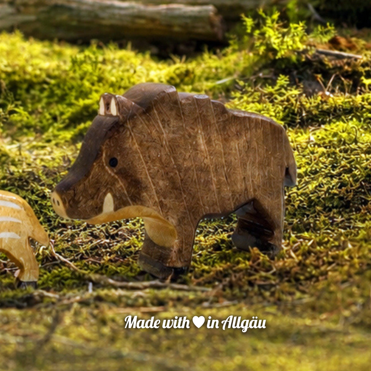 Handgefertigtes Holzspielzeug Wildschwein Bache von Allgäuer Holztiere, auf einem moosbedeckten Waldboden vor einem Baumstamm stehend, umgeben von natürlichen Waldmaterialien, im Hintergrund eine unscharfe Waldlandschaft, aus bayerischem Ahornholz gefertigt, mit pflanzlichen Farben braun, hellbraun und schwarz bemalt, im Allgäu produziert und für Kinder ab 3 Jahren geeignet.