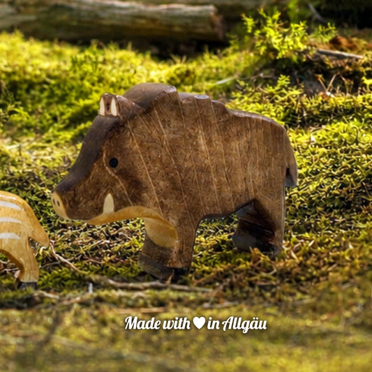 Handgefertigtes Holzspielzeug Wildschwein Bache von Allgäuer Holztiere, auf einem moosbedeckten Waldboden vor einem Baumstamm stehend, umgeben von natürlichen Waldmaterialien, im Hintergrund eine unscharfe Waldlandschaft, aus bayerischem Ahornholz gefertigt, mit pflanzlichen Farben braun, hellbraun und schwarz bemalt, im Allgäu produziert und für Kinder ab 3 Jahren geeignet.
