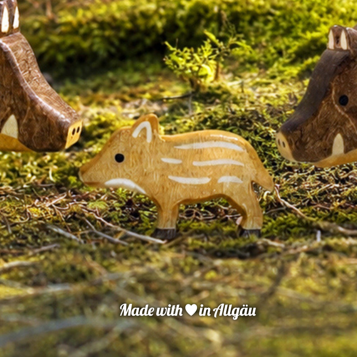 Handgefertigtes Holzspielzeug Wildschwein Frischling von Allgäuer Holztiere, auf einem moosbedeckten Waldboden zwischen einem Wildschwein-Keiler und einer Wildschwein-Bache stehend, umgeben von natürlichen Waldmaterialien, im Hintergrund eine unscharfe Waldlandschaft, aus bayerischem Ahornholz gefertigt, mit pflanzlichen Farben hellbraun und schwarz bemalt, im Allgäu produziert und für Kinder ab 3 Jahren geeignet.
