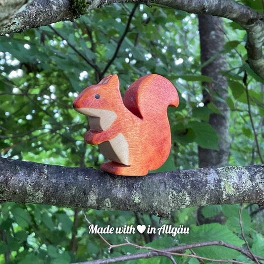 Handgefertigtes Holzspielzeug Eichhörnchen weiblich von Allgäuer Holztiere, braun bemalt mit pflanzlichen Farben, sitzend auf einem Ast in natürlicher Umgebung, aus bayerischem Ahornholz gefertigt, im Allgäu von Hand hergestellt und für Kinder ab 3 Jahren geeignet.