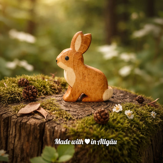 Handgefertigtes Holzspielzeug Feldhase  Baby von Allgäuer Holztiere, auf einem moosbedeckten Baumstumpf im Wald, umgeben von Tannenzapfen und kleinen weißen Blumen sitzend, im Hintergrund unscharfe Natur, aus bayerischem Ahornholz gefertigt, mit pflanzlichen Farben braun und schwarz bemalt, im Allgäu produziert und für Kinder ab 3 Jahren geeignet.